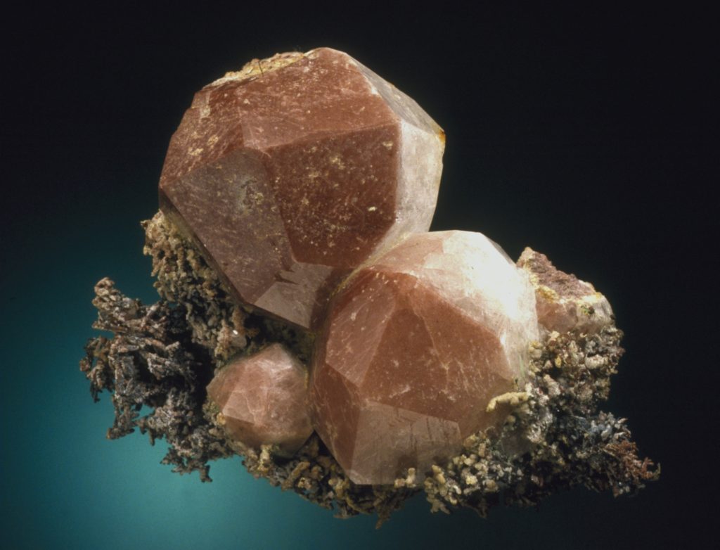 3 brownish-tan round crystals with many crystal faces on them, all sitting on microscopic copper