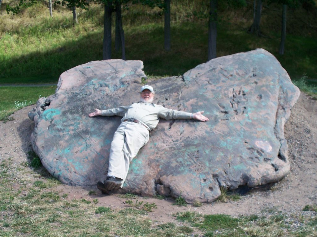 A very large piece of copper moved and smoothed by the glaciers with a man laying on top of it for scale. The copper makes the man look small. 